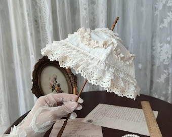 Könnte beinhalten: Ein kleiner weißer Spitzenschirm mit gestuften Rüschen und einem dünnen Holzgriff. Der Schirm wird von einer Hand mit weißem Spitzenhandschuh gehalten, neben einem Holzlineal, einem Blatt Papier im Vintage-Stil und einem Perlenarmband.