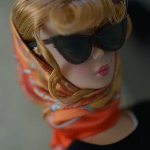 May include: A doll with blonde hair styled in a side-swept fringe, wearing black cat-eye sunglasses and an orange patterned headscarf tied under the chin. The doll has a beauty mark on the cheek, pink lips, and wears a black top.