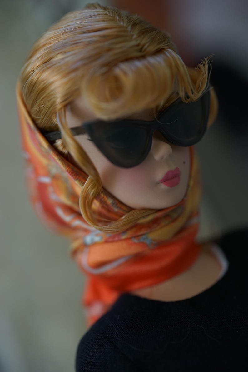 May include: A doll with blonde hair styled in a side-swept fringe, wearing black cat-eye sunglasses and an orange patterned headscarf tied under the chin. The doll has a beauty mark on the cheek, pink lips, and wears a black top.