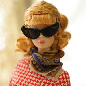 May include: A doll with blonde hair styled in a curled bob, wearing black cat-eye sunglasses, a patterned silk neck scarf in blue, gold, and white, and a red and white houndstooth print top. The doll has pink lips and a small beauty mark.