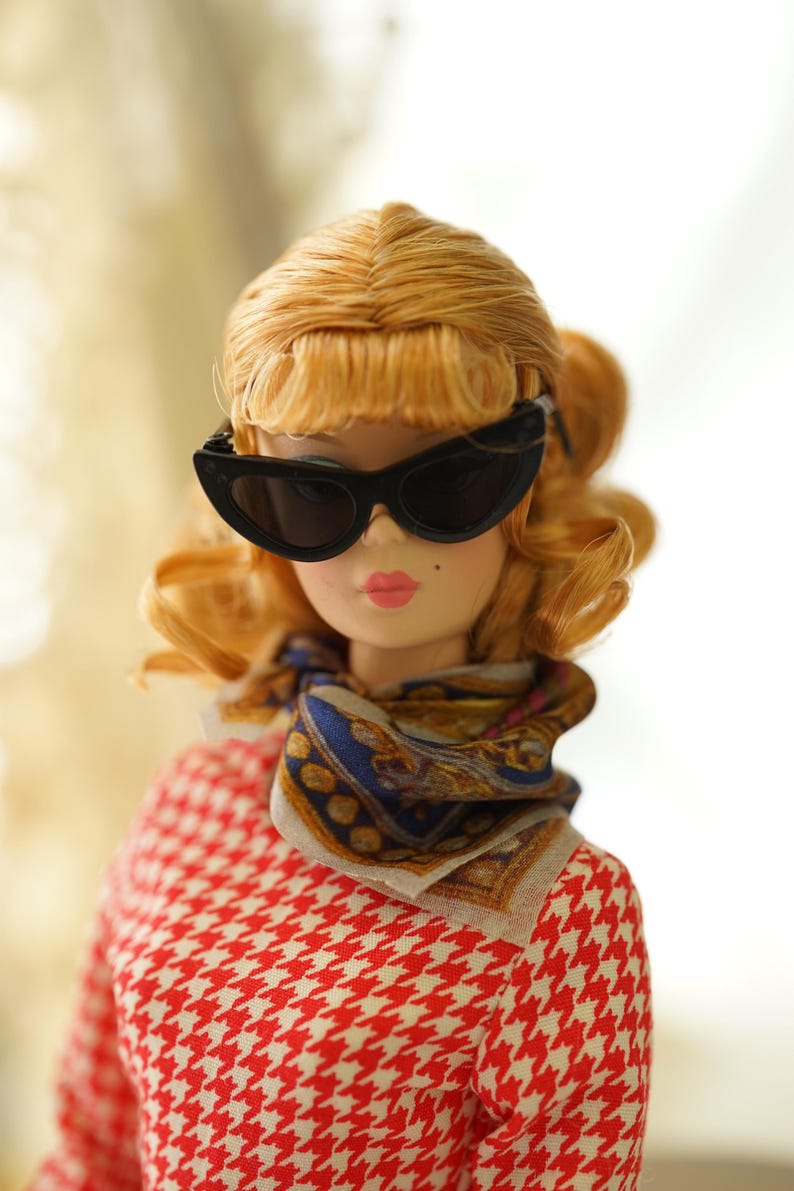 May include: A doll with blonde hair styled in a curled bob, wearing black cat-eye sunglasses, a patterned silk neck scarf in blue, gold, and white, and a red and white houndstooth print top. The doll has pink lips and a small beauty mark.