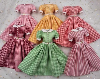 Könnte beinhalten: Sechs Puppenkleider mit kurzen Puffärmeln, weißen Bubikragen und weißen Taillengürteln mit goldenen Schnallen. Die Kleider sind in verschiedenen Vichy-Karomustern in Rosa, Gelb, Rot und Grün sowie einem Kleid mit rosa-weißen Streifen erhältlich.