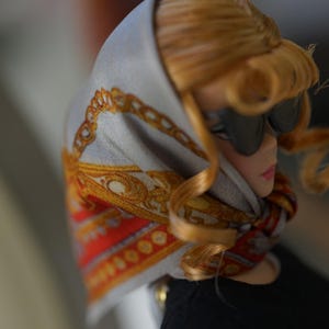 May include: A doll with blonde hair wearing black sunglasses and a light gray headscarf featuring a gold chain pattern and red accents. The doll is dressed in a black top, with the scarf tied around the neck, showcasing detailed fabric prints.