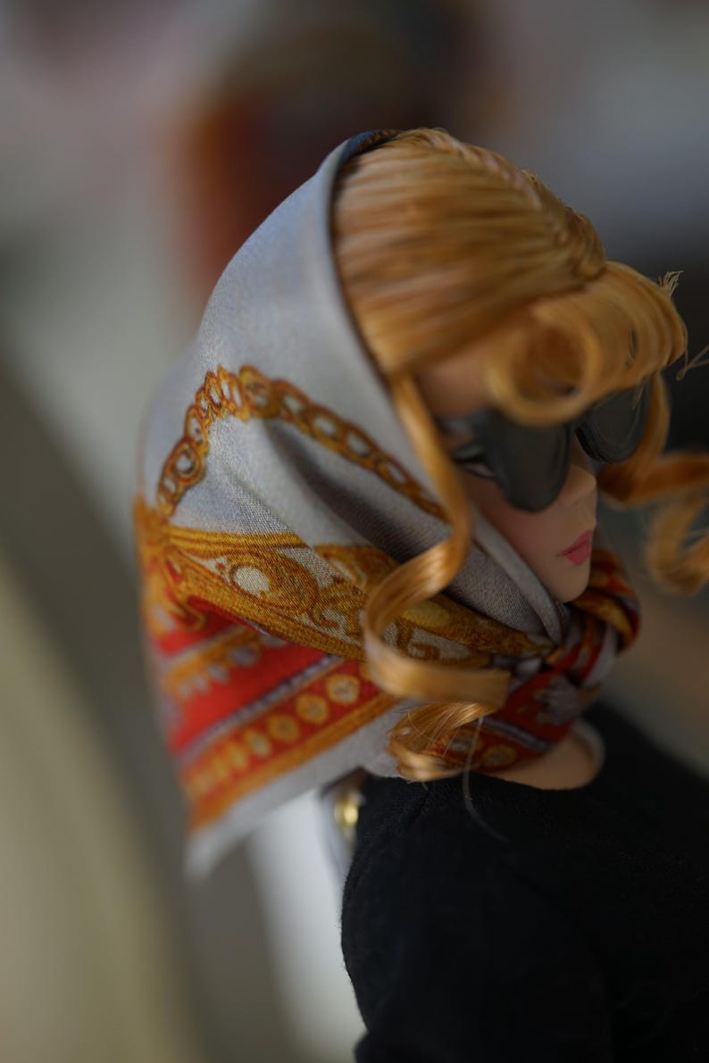 May include: A doll with blonde hair wearing black sunglasses and a light gray headscarf featuring a gold chain pattern and red accents. The doll is dressed in a black top, with the scarf tied around the neck, showcasing detailed fabric prints.