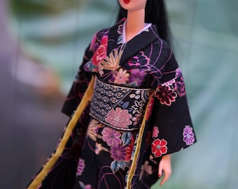 Könnte beinhalten: Eine Puppe mit langem, schwarzem Haar in einem hohen Pferdeschwanz, bekleidet mit einem schwarzen Kimono mit einem lebendigen Blumenmuster in Rosa-, Lila- und Weißtönen. Das Kleidungsstück hat einen breiten, strukturierten schwarzen Obi-Gürtel.