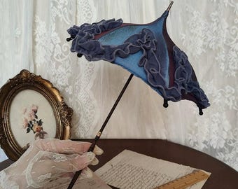 Könnte beinhalten: Ein Miniatur-Sonnenschirm mit einem dunkelblauen und hellblauen Stoffdesign, akzentuiert mit Rüschenbesatz. Der Griff ist dunkelbraun mit einer dekorativen Spitze. Der Sonnenschirm wird über einem Vintage-Dokument und einem Holzlineal gehalten.