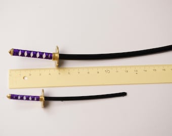 Könnte beinhalten: Zwei Miniatur-Spielzeug-Katanas mit lila und weiß umwickelten Griffen und goldfarbenen Parierstangen. Die schwarzen Klingen sind gebogen. Ein gelbes Lineal mit Zentimetermarkierungen liegt neben den Schwertern und zeigt ihre Größe.