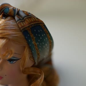 May include: A fabric headband featuring a knotted top detail with a printed pattern of teal, gold, and brown geometric shapes and small white dots. The headband is worn on a doll with light brown hair, blue eyes, and a small beauty mark on the cheek.