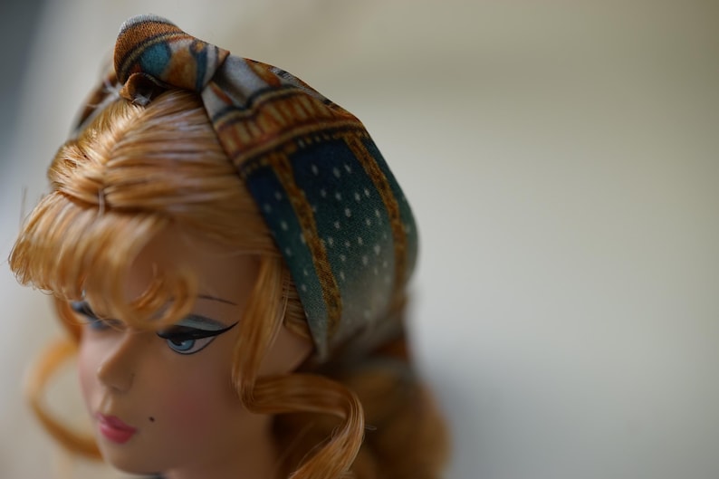 May include: A fabric headband featuring a knotted top detail with a printed pattern of teal, gold, and brown geometric shapes and small white dots. The headband is worn on a doll with light brown hair, blue eyes, and a small beauty mark on the cheek.