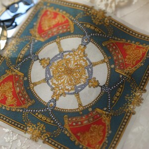 May include: A square silk scarf featuring a central gold filigree medallion surrounded by four red and gold shield-like emblems on a deep teal background. The design includes intricate chain-link patterns and a decorative border with gold dots.