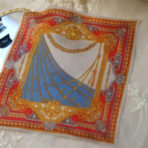May include: A square pocket square featuring a vibrant red border with gold filigree and chain motifs surrounding a central blue section with diagonal gold chain patterns. The design includes ornate gold frames and intricate decorative elements.