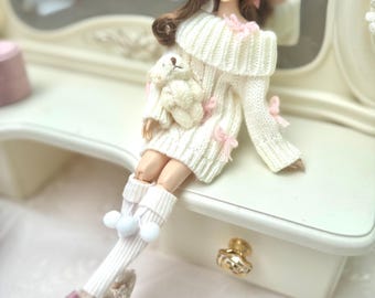 Könnte beinhalten: Eine Modepuppe mit langem braunem Haar und einer rosa Schleife, bekleidet mit einem schulterfreien, cremefarbenen Zopfmuster-Pullover mit kleinen rosa Schleifen, weißen gerippten Kniestrümpfen mit Bommeln und flauschigen Hausschuhen mit rosa Schleifen.