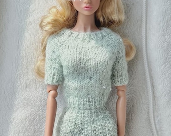 May include: A fashion doll with blonde hair wearing a light mint green knitted short-sleeved dress with a textured pattern and a matching pillbox hat featuring a delicate white netting veil that covers the upper portion of the doll's face.