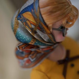 May include: A doll with light brown hair wearing a patterned headscarf featuring blue, orange, and gold geometric designs. The doll has dark-rimmed glasses and is dressed in a mustard yellow top with a small brown bow detail at the neckline.