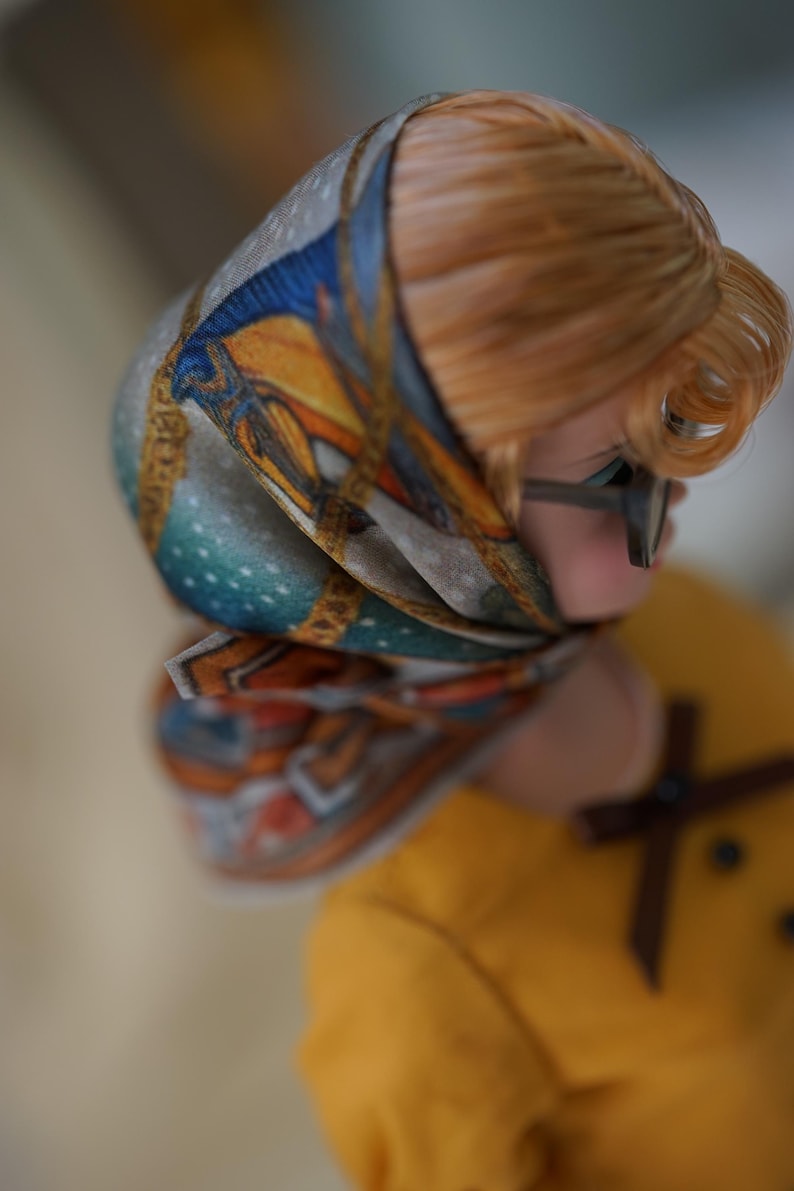 May include: A doll with light brown hair wearing a patterned headscarf featuring blue, orange, and gold geometric designs. The doll has dark-rimmed glasses and is dressed in a mustard yellow top with a small brown bow detail at the neckline.