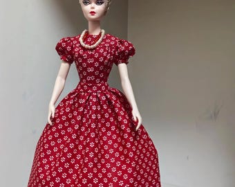 Könnte beinhalten: Eine Puppe mit blondem, hochgestecktem Haar, bekleidet mit einem roten Kleid mit weißem Blumenmuster, kurzen Puffärmeln und einem weiten Rock. Die Puppe trägt eine zweireihige Halskette mit weißen Perlen und kleinen roten Perlen.