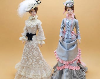 Könnte beinhalten: Zwei Puppen in aufwendigen Outfits. Die linke Puppe trägt ein cremefarbenes Spitzenkleid mit schwarzer Schleife und einen breitkrempigen Hut mit weißer Feder. Die rechte Puppe trägt ein graues Satinkleid mit rosa Rüschen und einen passenden Hut.