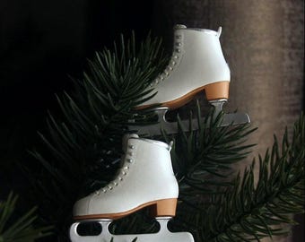 Könnte beinhalten: Zwei Miniatur-Eislaufschuhe in Weiß mit hellbraunen Absätzen und silberfarbenen Kufen. Jeder Schlittschuh hat ein hohes Design mit weißen Schnürsenkeln und silbernen Ösen, platziert zwischen dunkelgrünen Tannennadeln.