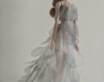 May include: A fashion doll with long auburn hair wearing a floor-length, strapless gown made of layered, translucent grey and white petal-like fabric. The bodice features delicate silver bead embellishments and thin, draped beaded shoulder straps.