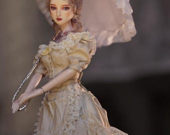 Könnte beinhalten: Eine Gelenkpuppe mit hellbraunem Haar, bekleidet mit einem cremefarbenen Kleid mit Spitzenbesatz, einer gerüschten Schürze und einer Halskette. Die Puppe hält einen weißen, gerüschten Sonnenschirm mit silbernem Griff. Der Text "soul factory" ist zu sehen.