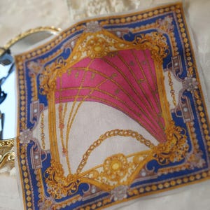 May include: A square silk scarf featuring a vibrant pink center with gold chain motifs, framed by a deep blue border with gold geometric patterns, positioned next to a gold-toned handheld mirror with black sunglasses resting on its reflective surface.