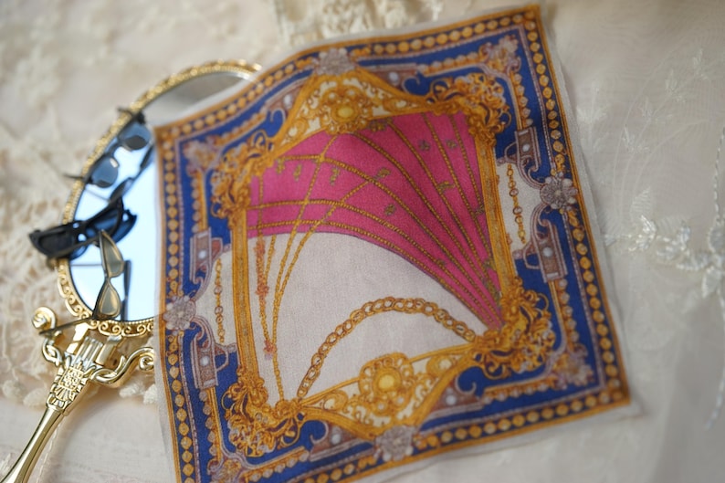 May include: A square silk scarf featuring a vibrant pink center with gold chain motifs, framed by a deep blue border with gold geometric patterns, positioned next to a gold-toned handheld mirror with black sunglasses resting on its reflective surface.