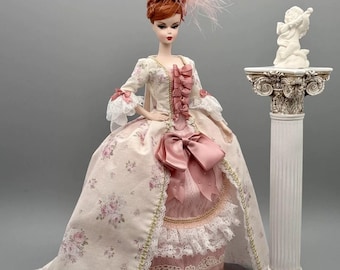 Könnte beinhalten: Eine Puppe mit rotem Haar in einer Hochsteckfrisur mit rosa Feder-Accessoire, bekleidet mit einem cremefarbenen Kleid mit rosa Blumenmuster, Spitzenbesatz und einer großen rosa Schleife an der Taille, neben einer weißen Säule mit einem Putto.