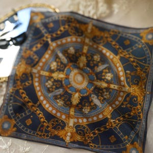 May include: A square silk scarf featuring a dark blue background with a gold and white ornate chain and medallion pattern. The design includes symmetrical circular and geometric motifs arranged in a central composition with intricate gold detailing.