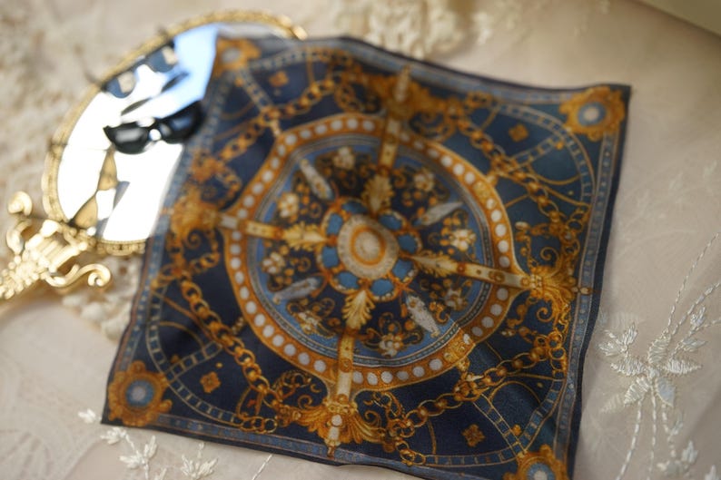 May include: A square silk scarf featuring a dark blue background with a gold and white ornate chain and medallion pattern. The design includes symmetrical circular and geometric motifs arranged in a central composition with intricate gold detailing.