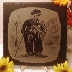 May include: A black slate plaque with a sepia-toned illustration of Charlie Chaplin. The image shows Chaplin in his signature outfit, including a bowler hat and suit, standing on a street with a cane. The background depicts a vintage street scene.