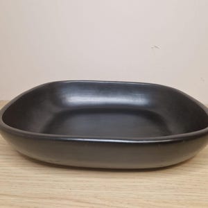 Op de afbeelding: Een zwarte, ovaalvormige keramische kom met afgeronde randen. De kom staat op een lichtgekleurd houten oppervlak. De binnenkant van de kom is glad en reflecteert licht. Het is een decoratief item.