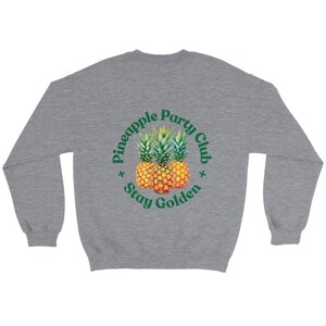 Puede incluir: Una sudadera gris jaspeado con un dise&ntilde;o circular que presenta tres pi&ntilde;as y el texto "Pineapple Party Club Stay Golden" en verde. Las pi&ntilde;as son amarillas y naranjas con copas verdes.