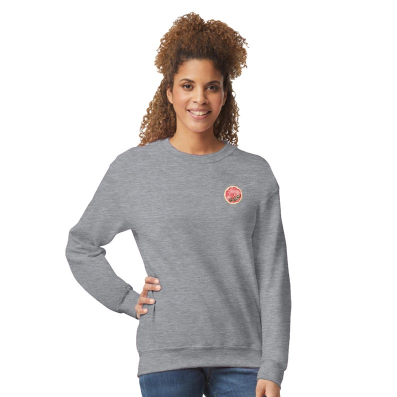 May include: A heather gray sweatshirt with a small, circular graphic on the upper left chest. The graphic features a pink and red design with a tan border. The sweatshirt has long sleeves and a crew neck.