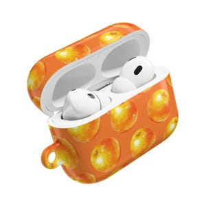 Könnte beinhalten: Orangenes AirPods-Case mit einem Aquarell-Fruchtmuster. Das Case ist geöffnet und zeigt weiße AirPods. Das Case hat eine kleine Schlaufe an der Seite. Der Hintergrund ist weiß.