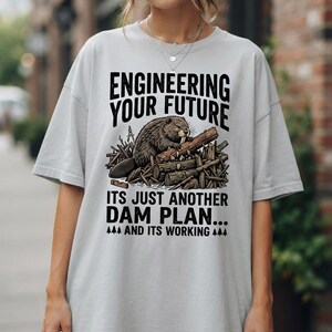 Puede incluir: Una camiseta gris claro con un gráfico de un castor sobre una presa y el texto "ENGINEERING YOUR FUTURE", "ITS JUST ANOTHER DAM PLAN..." y "AND ITS WORKING", acompañado de pequeños iconos de pinos e ilustraciones de herramientas de dibujo.