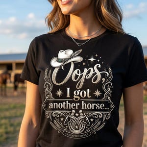 Op de afbeelding: Een zwart t-shirt met een grafische print van een witte cowboyhoed, hoefijzers en korenaren. De tekst luidt: "Oops, I got another horse..." en "A LIFESTYLE CHOICE." Het ontwerp bevat decoratieve krullen en sterren in een crèmekleurige tint.