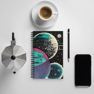 May include: Spiral-bound notebook featuring a colorful space-themed design with planets in teal, pink, and white against a black background. A cup of coffee, a pencil, a phone, and a coffee maker are also visible.