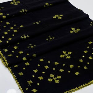 Peut inclure: Tissu noir orné de broderies florales et en croix de couleur jaune doré. Les bords sont finis avec une bordure au crochet jaune doré assortie. Le tissu est plié, révélant la broderie et la texture.