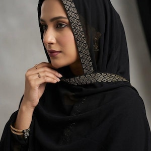 May include: A black head covering with a gold-colored trim. The trim is decorated with small, evenly spaced dots. The fabric drapes over the shoulders. A gold bracelet and ring are visible.