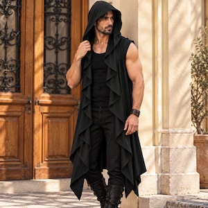 May include: A black sleeveless hooded vest featuring a draped, ruffled open front design that extends to a longer length. The garment is worn over a black tank top, black trousers, and black boots, creating a layered, monochromatic appearance.