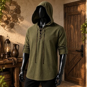 May include: An olive green hooded shirt featuring long sleeves with roll-up tabs, a lace-up front closure with thin brown cords, and a curved hemline. The garment is displayed on a black mannequin, showcasing a textured fabric construction.