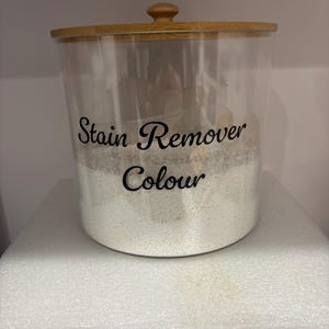 May include: A transparent, cylindrical container with a wooden lid and knob, labelled "Stain Remover Colour" in black script. The container is filled with a white powder, likely a laundry product.
