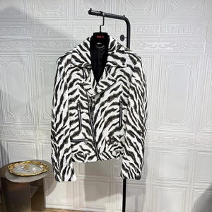 May include: A white and black zebra print jacket with a black collar and zippers. The jacket is hanging on a black clothing rack. The jacket has a classic biker jacket design.