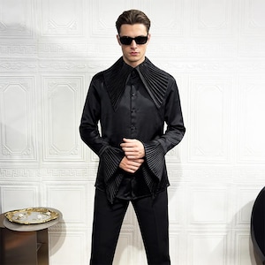 Puede incluir: Camisa negra de manga larga con cuello y puños plisados. La camisa tiene una botonadura frontal y se combina con pantalones negros. El modelo lleva gafas de sol. El diseño de la camisa es una versión única de la ropa formal.