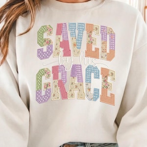 Könnte beinhalten: Cremefarbenes Sweatshirt mit dem Text "SAVED by His GRACE" in einem Patchwork-Design. Die Buchstaben sind mit Blumen- und Tupfenmustern in Pastellfarben gefüllt, was eine weiche, Vintage-Ästhetik erzeugt.