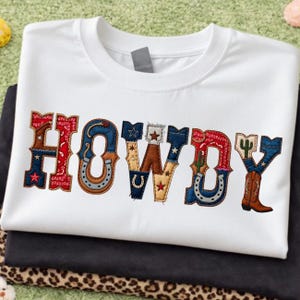 May include: White t-shirt with the word "HOWDY" in a colorful, Western-themed design. The letters are decorated with stars, horseshoes, and cowboy boot illustrations. The shirt is folded on top of black fabric.