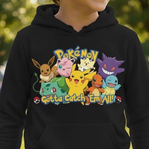 May include: Black hooded sweatshirt featuring a colorful graphic of various Pokémon characters, including Pikachu, Charmander, and Squirtle. The text "Pokémon" is above the characters, and "Gotta Catch 'Em All!" is below.