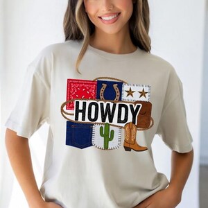 Może przedstawiać: Kremowa koszulka z grafiką "HOWDY" i elementami w stylu kowbojskim. Projekt zawiera czerwoną chustę, podkowę, gwiazdy, brązowy but, kaktus i naszywki z dżinsu, wszystko połączone liną.