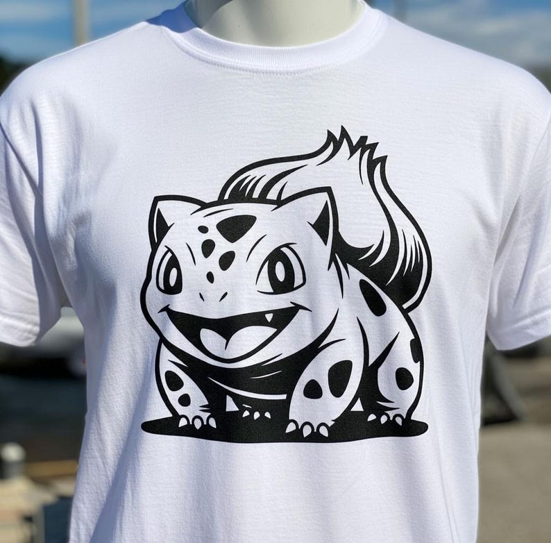 May include: White t-shirt featuring a black and white graphic of a cartoon character with a smiling face and spotted body. The design is centered on the front of the shirt. The shirt is displayed on a mannequin.