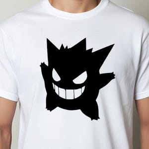 May include: White t-shirt featuring a black silhouette of a cartoon character with a jagged head, large eyes, and a wide grin. The design is centered on the shirt.